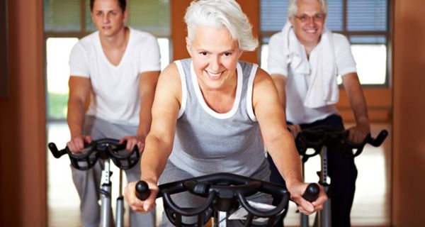 Fitte Seniorin mit grauen Kurzhaarschnitt im Fitnessstudio (Frontalbild) auf dem Fahrrad im Hintergrund zwei Männer