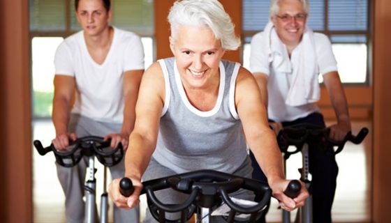 Fitte Seniorin mit grauen Kurzhaarschnitt im Fitnessstudio (Frontalbild) auf dem Fahrrad im Hintergrund zwei Männer