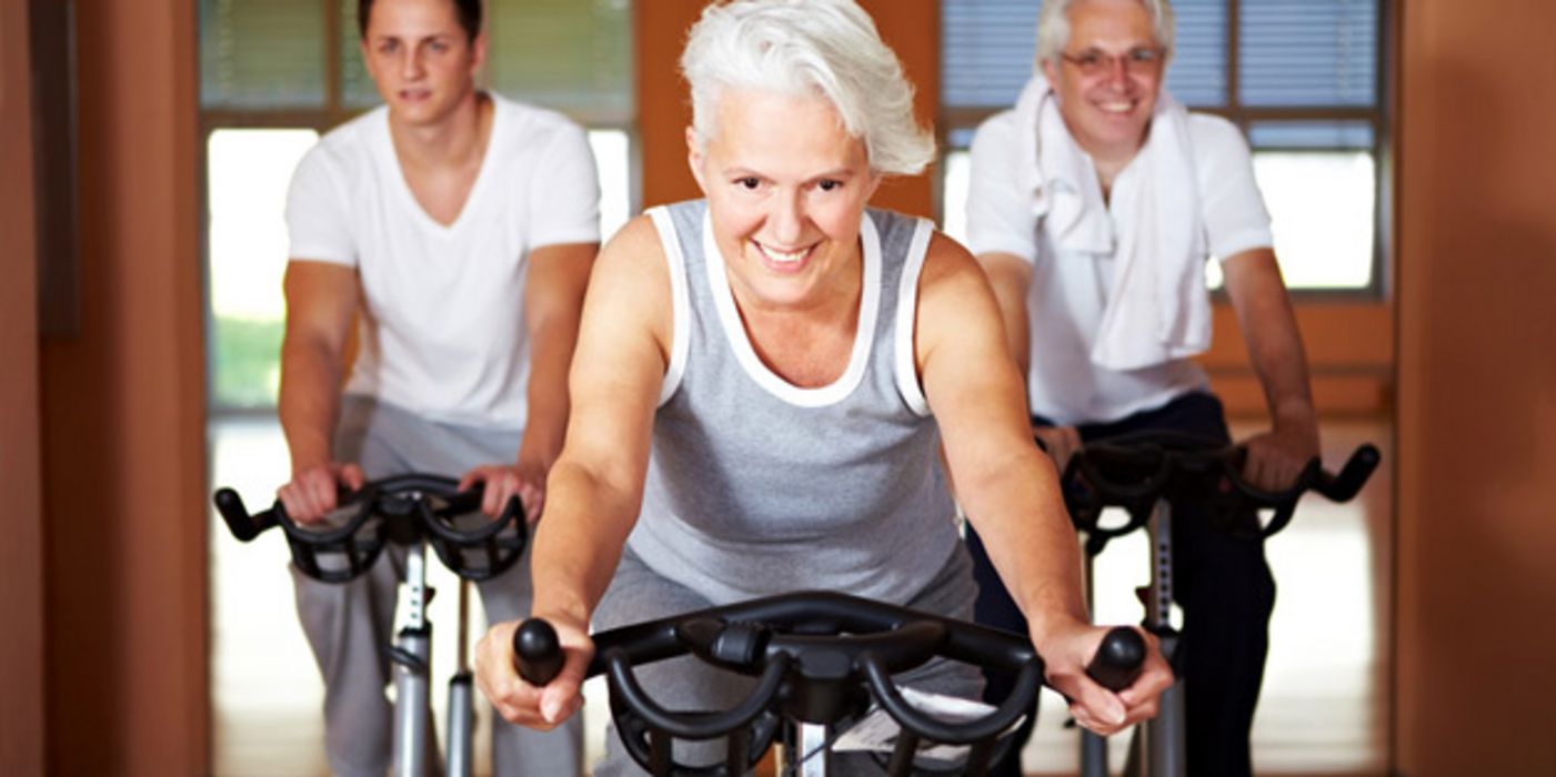 Fitte Seniorin mit grauen Kurzhaarschnitt im Fitnessstudio (Frontalbild) auf dem Fahrrad im Hintergrund zwei Männer