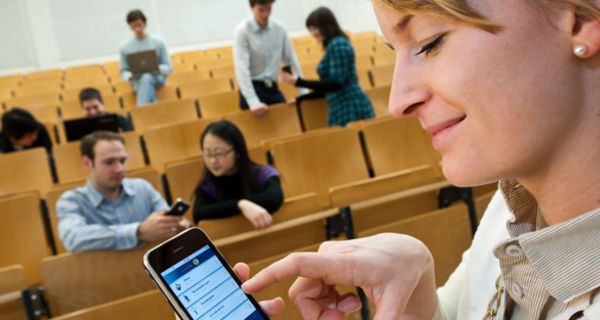 Hörsaal im Hintergrund mit Studierenden, Profil einer Studentin im Vordergrund die auf ihrem Smartphone tippt