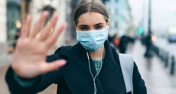 Frau mit Maske, hält eine Hand in die Kamera.