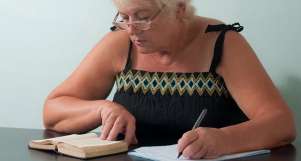 Frau um die 60 mit Brille schreibt etwas aus einem Buch ab