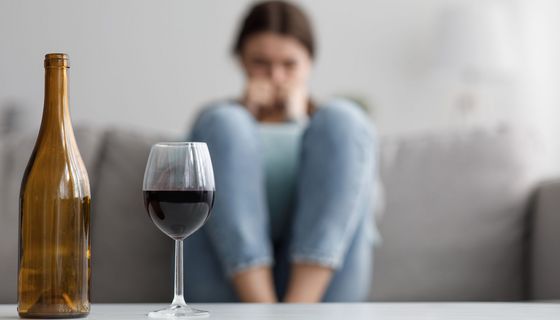 Ein Weinglas und eine leere Weinflasche stehen auf einem Tisch, während im Hintergrund eine junge Frau mit angezogenen Beinen auf einem Sofa sitzt.
