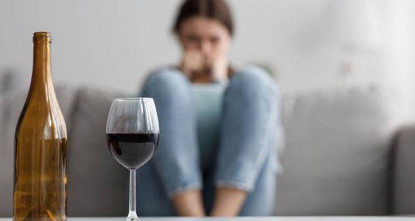 Ein Weinglas und eine leere Weinflasche stehen auf einem Tisch, während im Hintergrund eine junge Frau mit angezogenen Beinen auf einem Sofa sitzt.