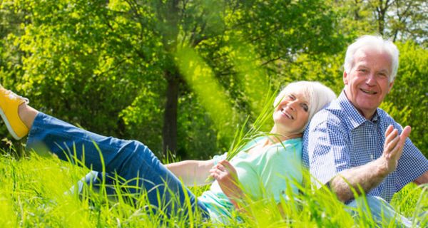 Rentnerpaar sitzt Rücken an Rücken auf einer Parkwiese und amüsiert sich; im Hintergrund Bäume