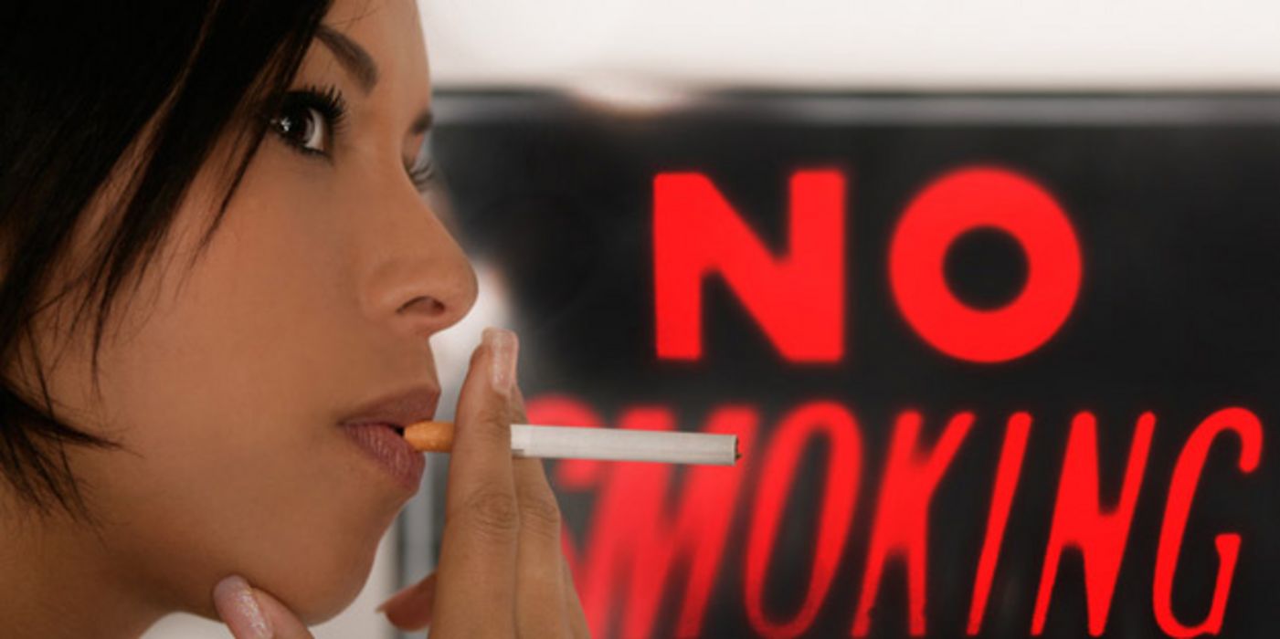 Portrait junge dunkelhaarige Frau im Profil mit Zigarette im Mund vor rotem Plakat "No Smoking"
