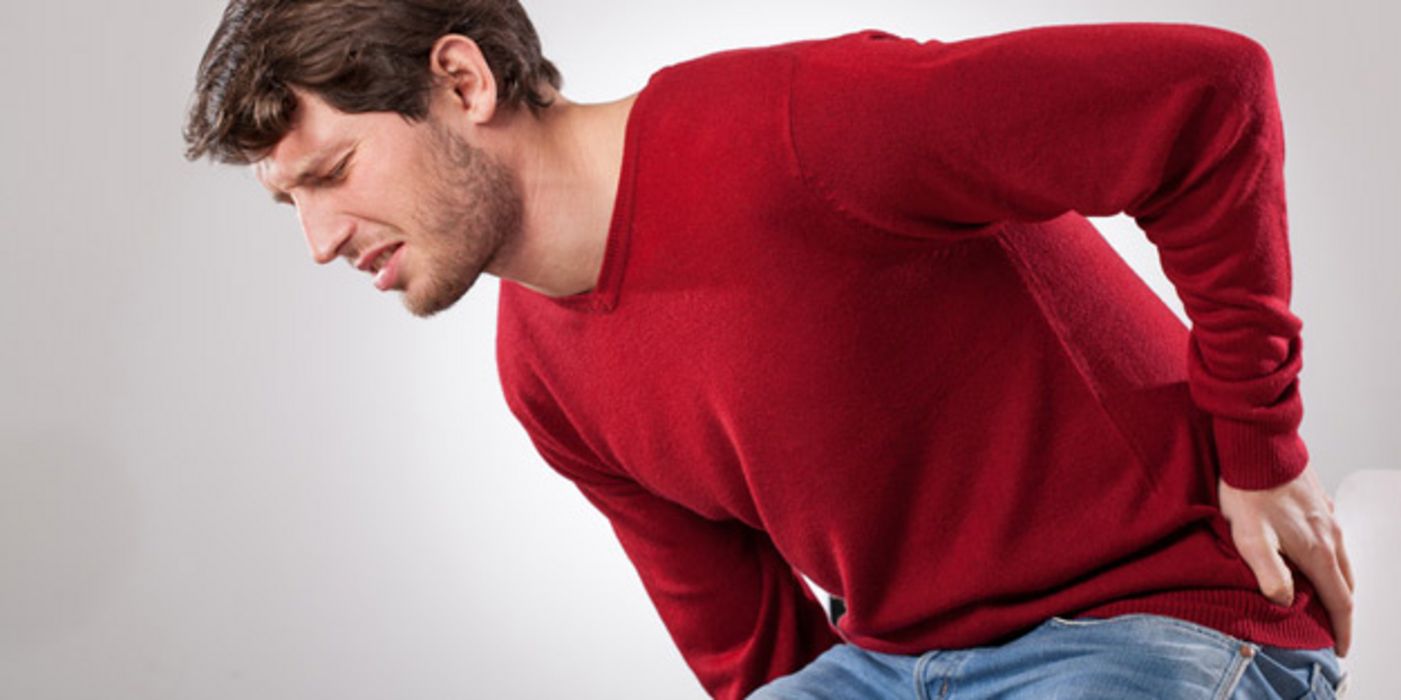 Dunkelhaariger Mann um die 30, Jeans, dunkelroter Pullover, hält sich mit schmerzverzerrtem Gesicht den unteren Rücken