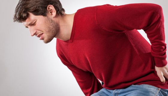 Dunkelhaariger Mann um die 30, Jeans, dunkelroter Pullover, hält sich mit schmerzverzerrtem Gesicht den unteren Rücken