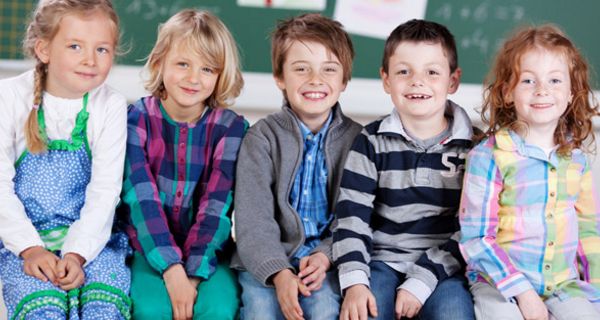 GrundschülerInnen in bunter Kleidung fröhlich lächelnd vor Tafel sitzend (2 Mädchen, 2 Jungen, 1 Mädchen, von links)
