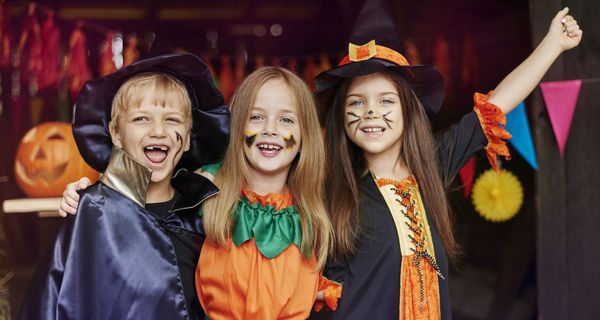 Ein Junge und zwei Mädchen – alle drei verkleidet als Zauberer, Kürbis und Hexe – am Eingang zu einer Halloween-Feier.