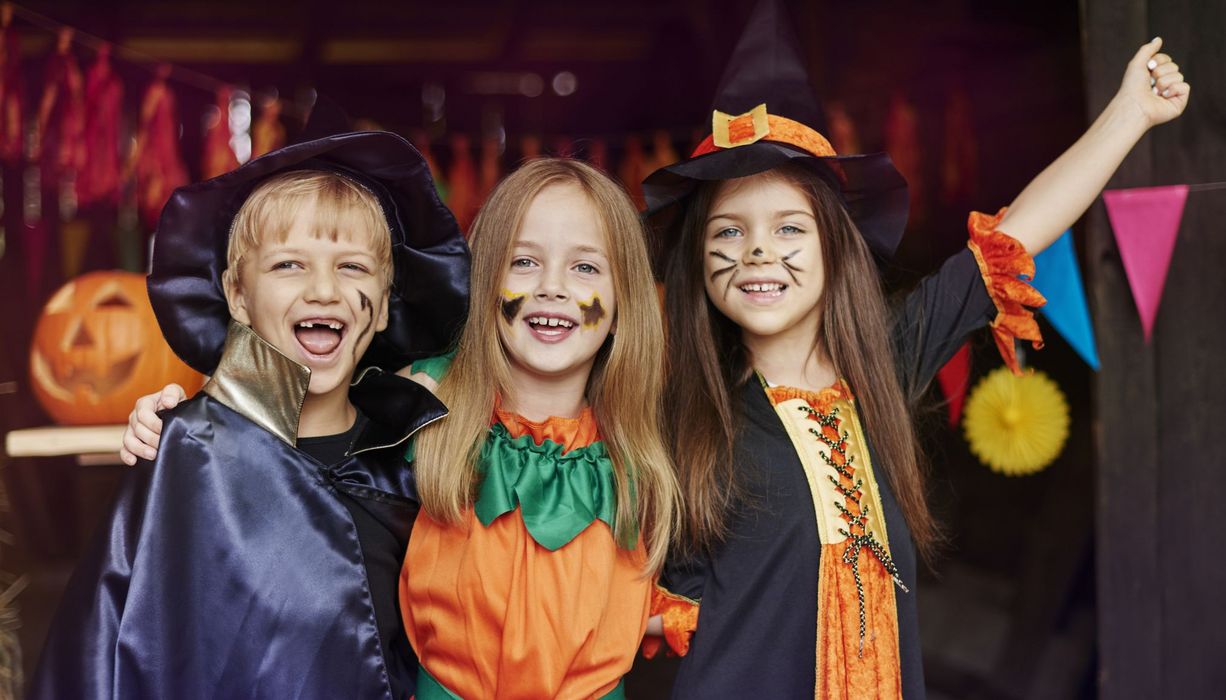 Ein Junge und zwei Mädchen – alle drei verkleidet als Zauberer, Kürbis und Hexe – am Eingang zu einer Halloween-Feier.