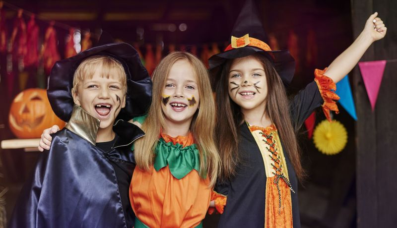Ein Junge und zwei Mädchen – alle drei verkleidet als Zauberer, Kürbis und Hexe – am Eingang zu einer Halloween-Feier.