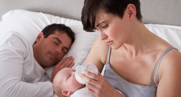 Junge Mutter im Bett sitzend schaut besorgt und gibt Baby ein Fläschchen während Mann schläft