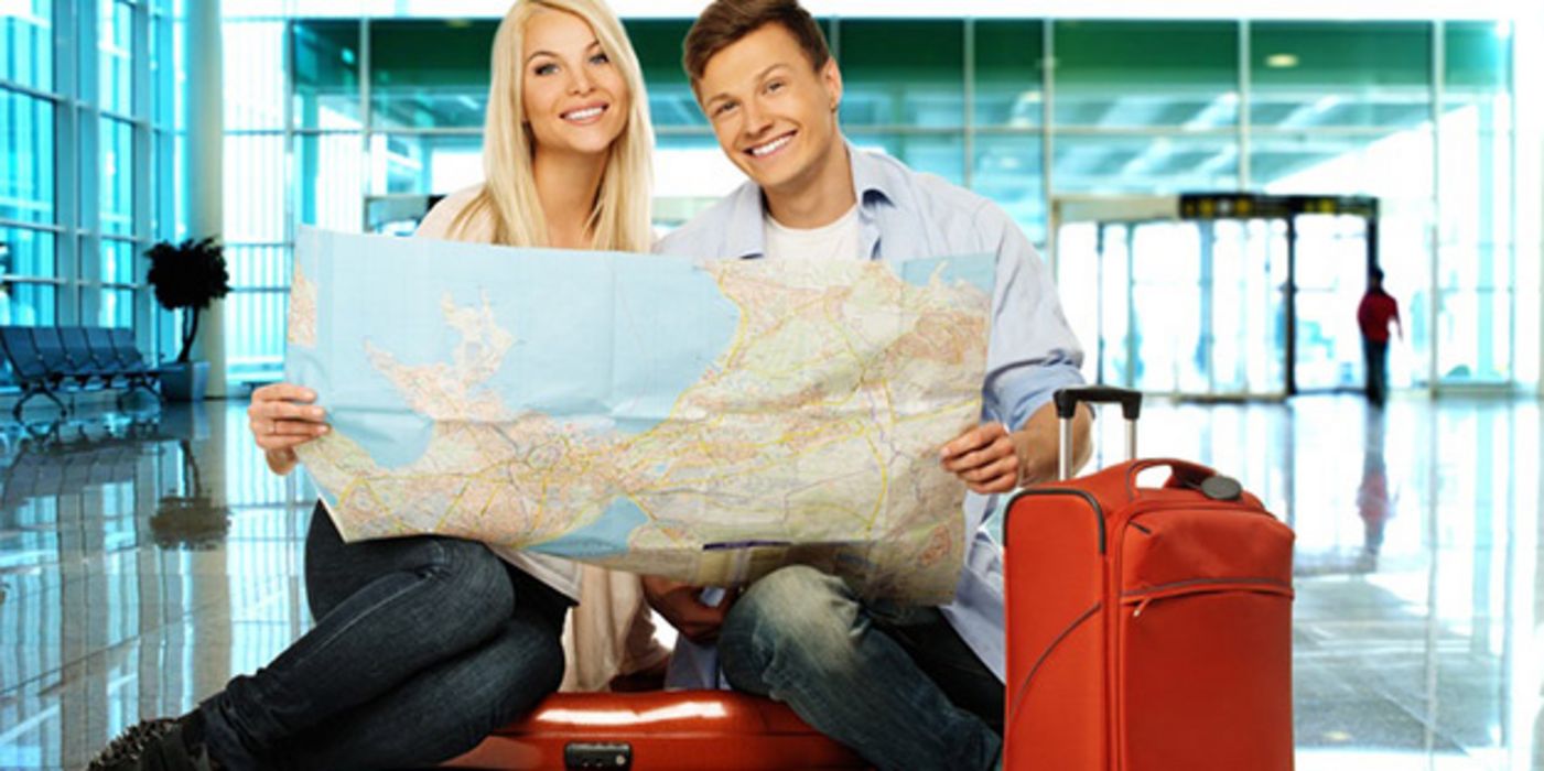 Frau und Mann, lachend, ca. 30+, Sitzen auf rotem Koffer in Flughafenabflughalle, roter Koffer neben Mann, und halten Landkarte in den Händen