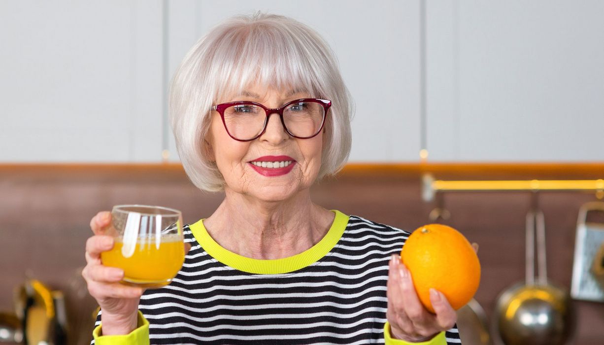 Eine ältere Dame hält ein Glas mit Orangen-Saft und eine Orange in der Hand.