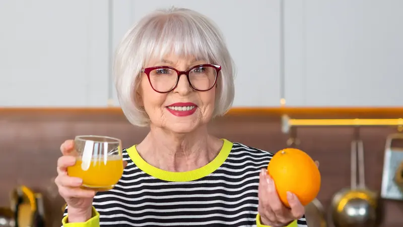 Eine ältere Dame hält ein Glas mit Orangen-Saft und eine Orange in der Hand.