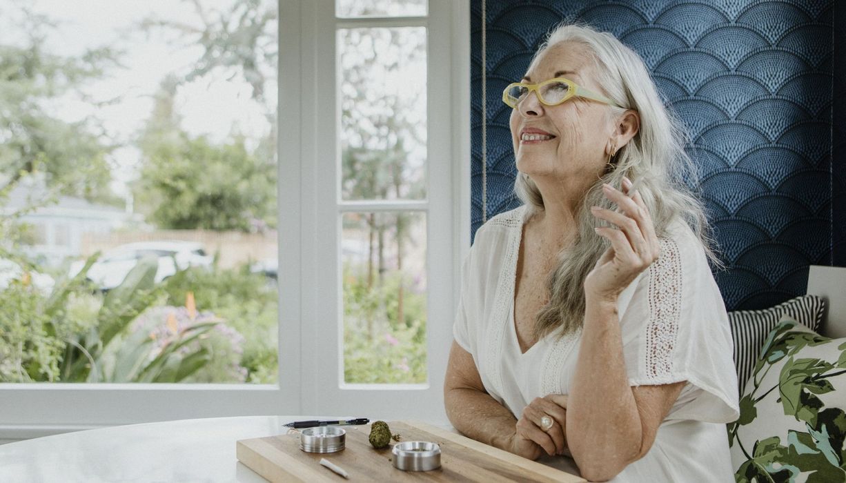 Seniorin sitzt im Bett und raucht einen Joint. Vor ihr liegt auf einem Brettchen ein weiterer sowie ein Bollen Cannabis.