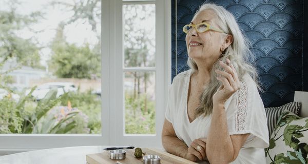 Seniorin sitzt im Bett und raucht einen Joint. Vor ihr liegt auf einem Brettchen ein weiterer sowie ein Bollen Cannabis.