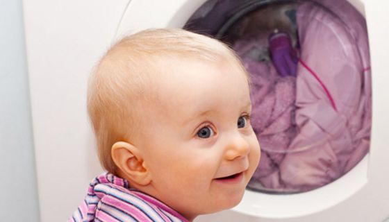 Großaufnahme runder Kopf und Schultern Kleinkind mit rosagestreiftem Oberteil vor Waschmaschinenauge mit Wäsche