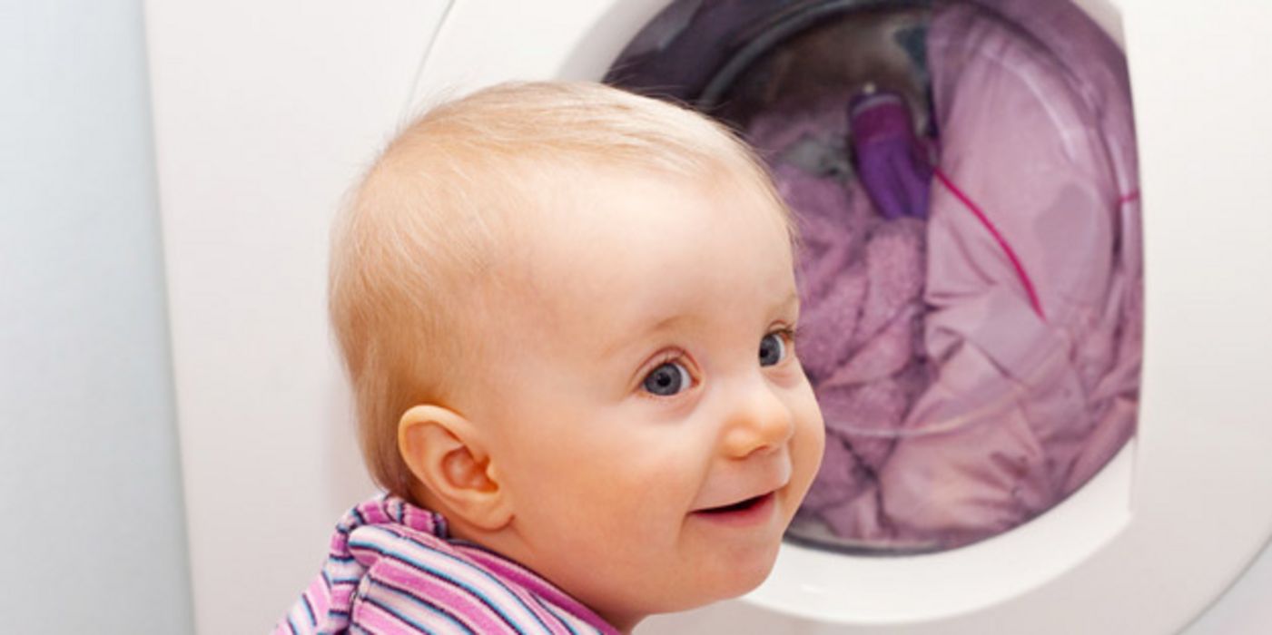 Großaufnahme runder Kopf und Schultern Kleinkind mit rosagestreiftem Oberteil vor Waschmaschinenauge mit Wäsche
