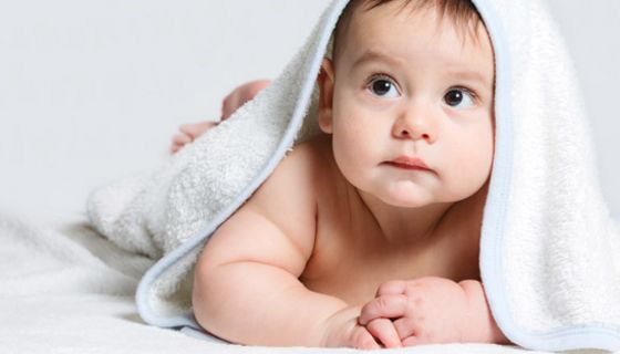 Properes Baby bäuchlings auf weißer Decke mit weißem Badetuch über dem Kopf, nach links oben blickend