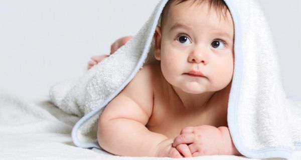 Properes Baby bäuchlings auf weißer Decke mit weißem Badetuch über dem Kopf, nach links oben blickend