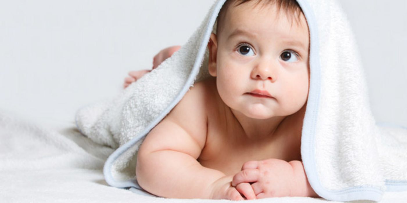 Properes Baby bäuchlings auf weißer Decke mit weißem Badetuch über dem Kopf, nach links oben blickend