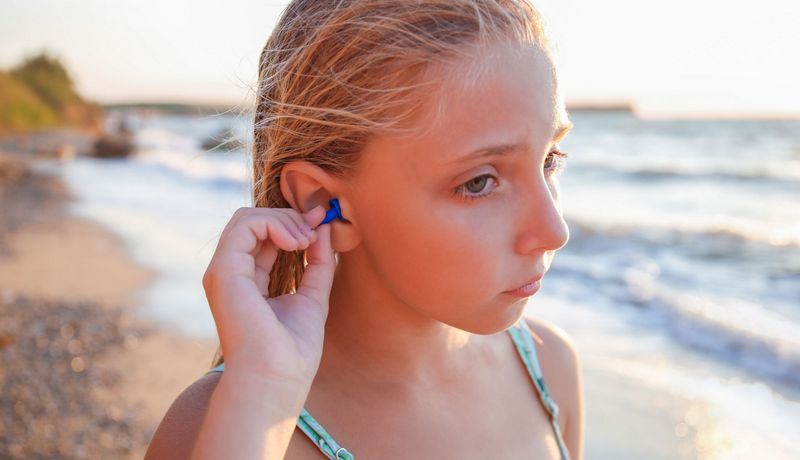 Mädchen, steht an einem Strand und steckt sich einen Schwimmschutz in die Ohren.