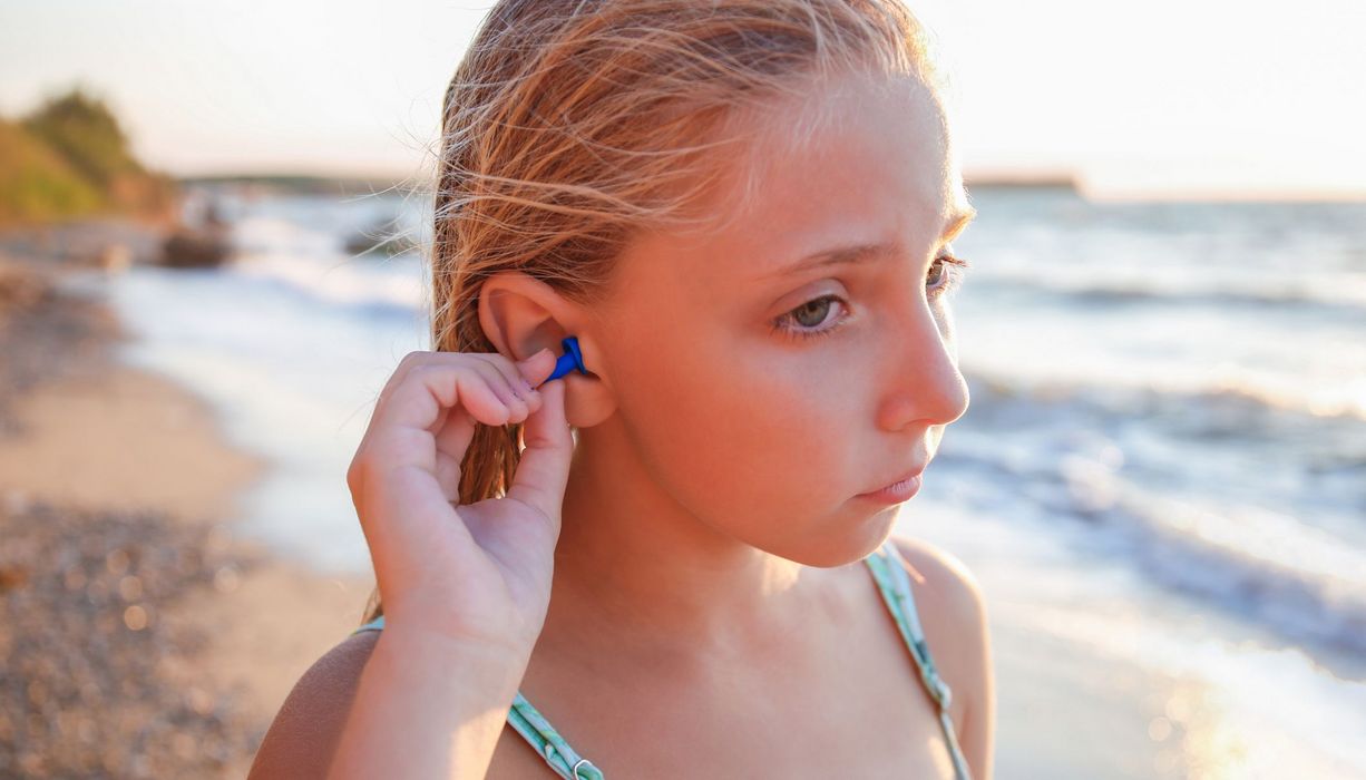 Mädchen, steht an einem Strand und steckt sich einen Schwimmschutz in die Ohren.