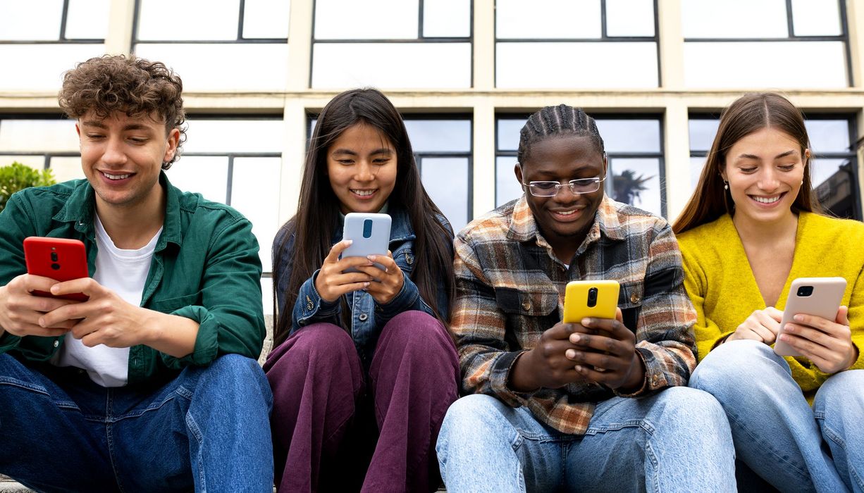 Vier Teenager sitzen in der Reihe vor einem Gebäude. Jeder hat ein Smartphone in der Hand in das er hineinschaut.
