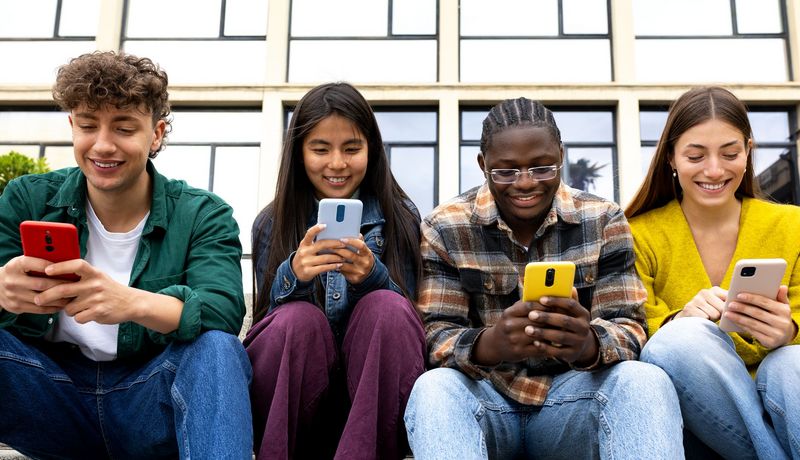 Vier Teenager sitzen in der Reihe vor einem Gebäude. Jeder hat ein Smartphone in der Hand in das er hineinschaut.