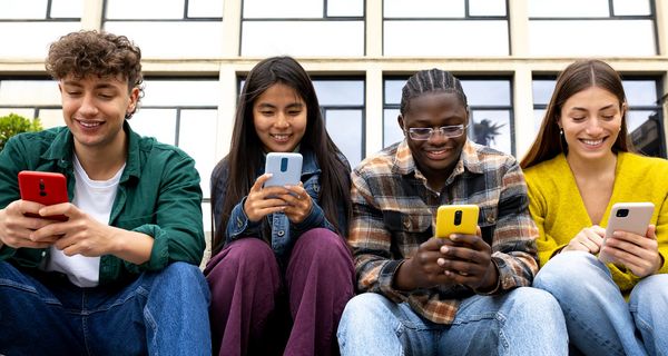 Vier Teenager sitzen in der Reihe vor einem Gebäude. Jeder hat ein Smartphone in der Hand in das er hineinschaut.