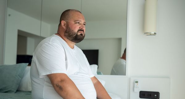 Beleibter Mann sitzt auf einem Bett.