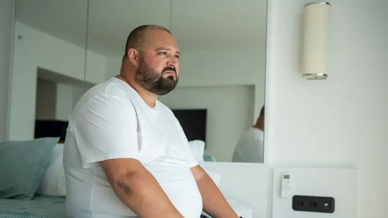 Beleibter Mann sitzt auf einem Bett.