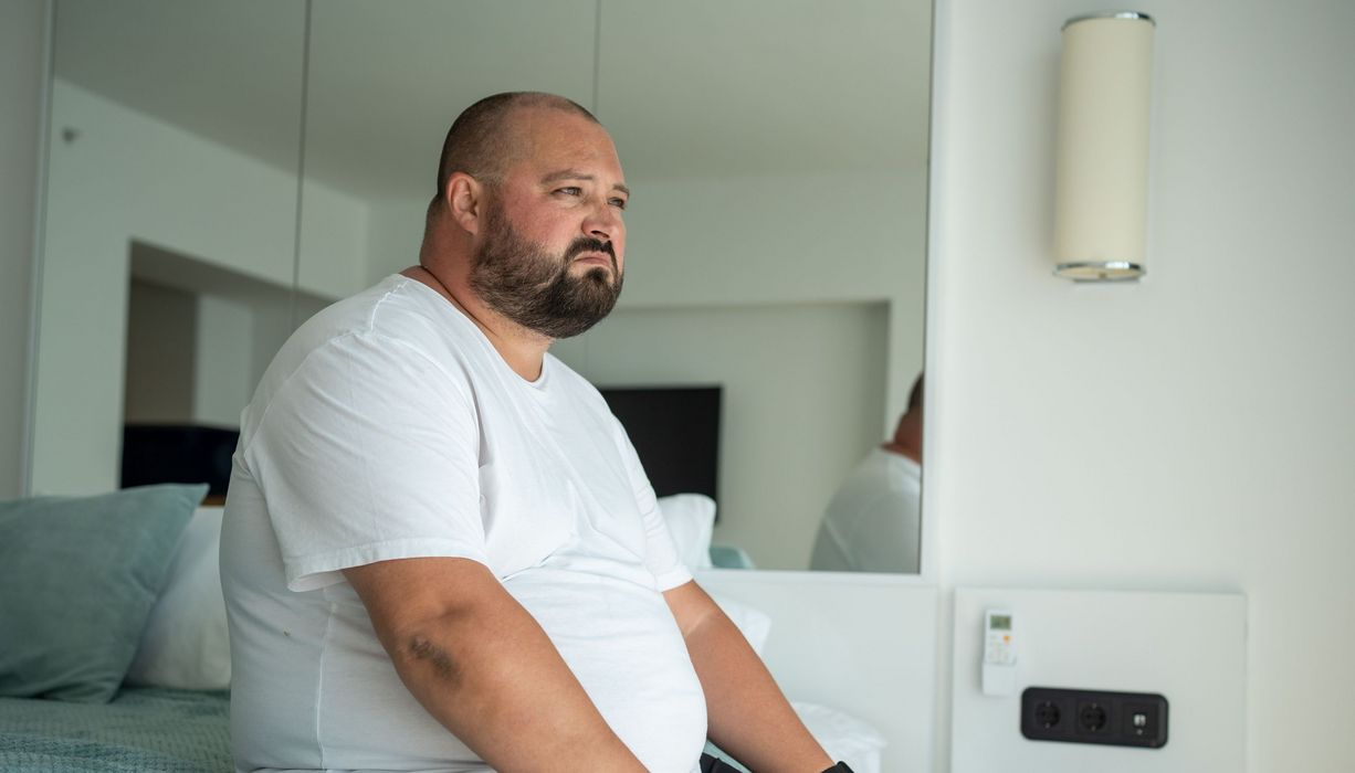 Beleibter Mann sitzt auf einem Bett.