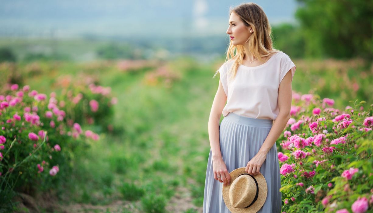 Junge Frau in weiter Bluser und Rock mit Sommerhut steht in einem Rosengarten.