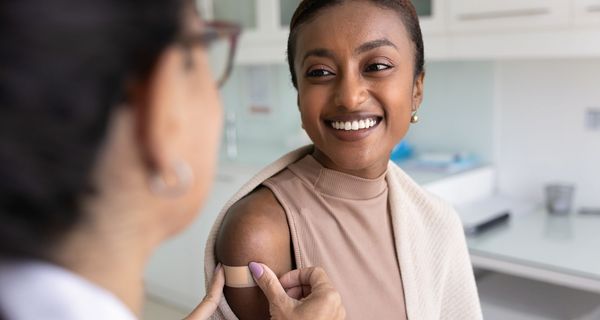 Eine Ärztin klebt einer lächelnden Frau ein Pflaster auf den Oberarm.