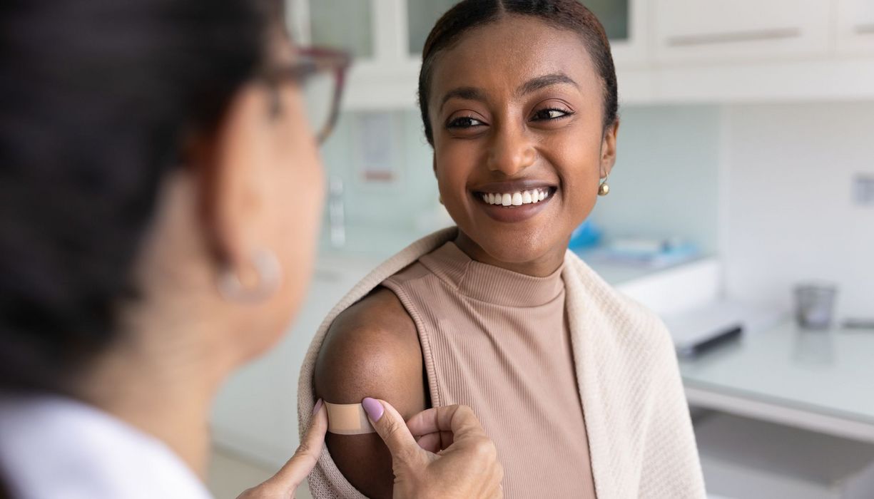 Eine Ärztin klebt einer lächelnden Frau ein Pflaster auf den Oberarm.