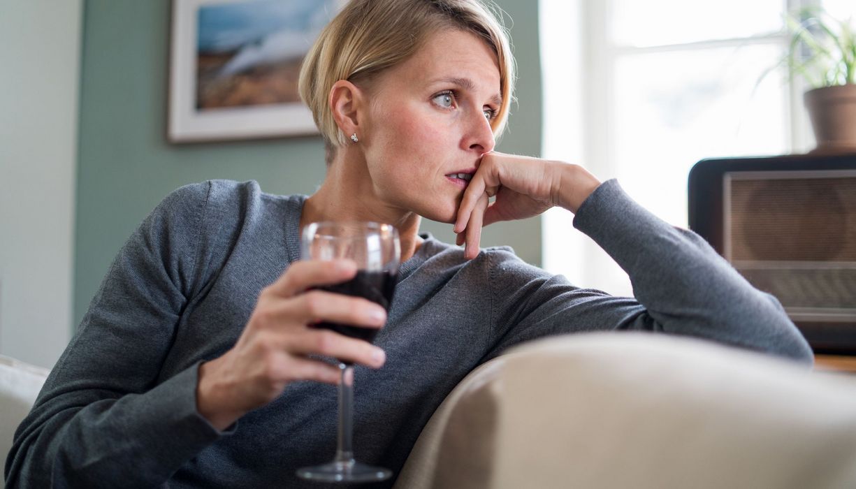 Frau sitzt am Tag mit einem Glas Rotwein auf dem Sofa und guckt nachdenklich.