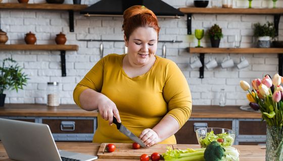 Eine junge, übergewichtige Frau steht in der Küche und schneidet Tomaten.