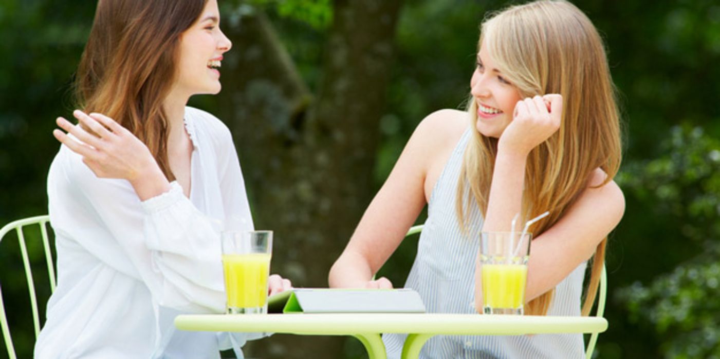 Zwei langhaarige Teenagerfrauen sitzen an einem Bistrotischchen im Freien, unterhalten sich und trinken O-Saft, im Hintergrund Wald