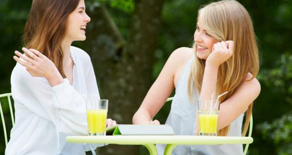 Zwei langhaarige Teenagerfrauen sitzen an einem Bistrotischchen im Freien, unterhalten sich und trinken O-Saft, im Hintergrund Wald