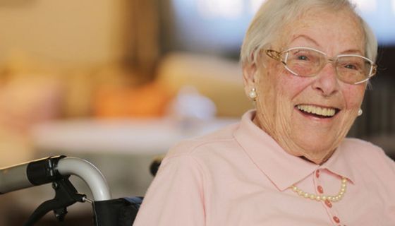 Lachende Seniorin im Rollstuhl sitzend, rosa Polopulli, graue, kurze Haare, Perlenohringe, -kett und schicke Brille 