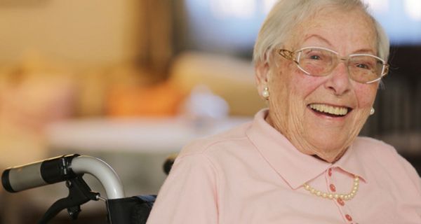 Lachende Seniorin im Rollstuhl sitzend, rosa Polopulli, graue, kurze Haare, Perlenohringe, -kett und schicke Brille 