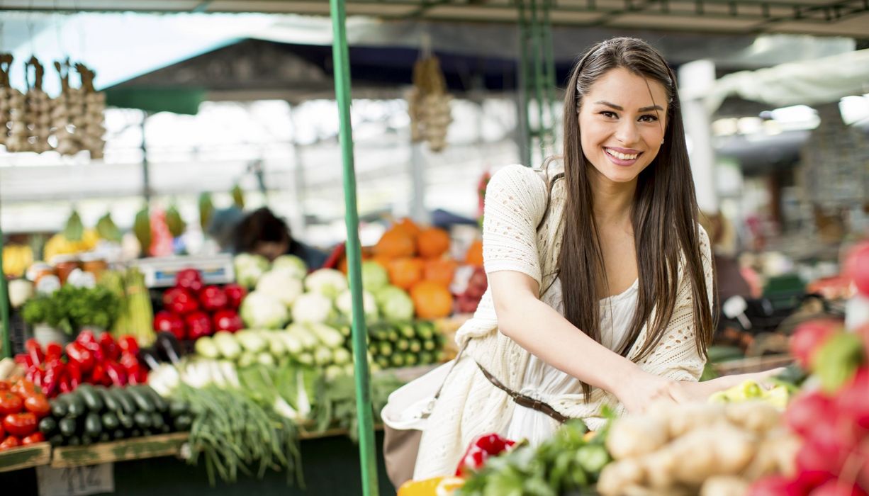 Junge Frau betrachtet einen Marktstand mit Obst und Gemüse.