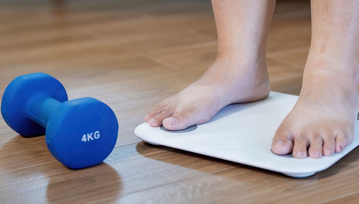 Zu sehen sind nur die Füßer einer Frau auf einer Waage, daneben eine 4kg-Hantel.