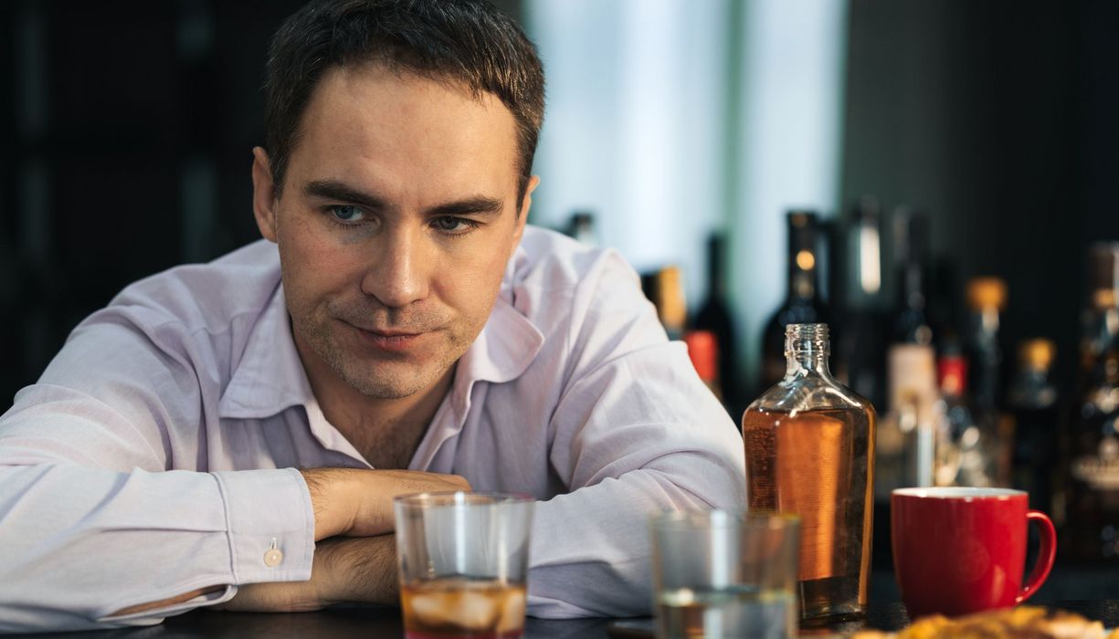 Mann schaut nachdenklich, vor und neben ihm stehen Gläser und Flaschen mit Alkohol. 