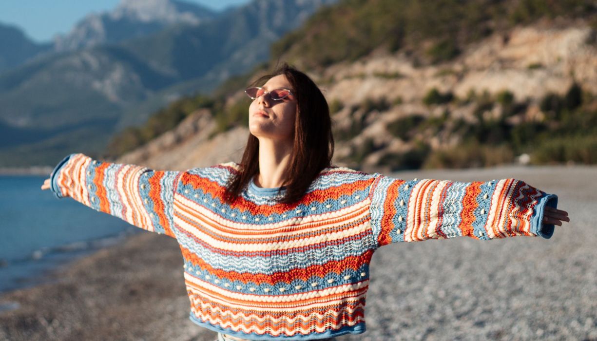 Junge Frau im bunten Pulli badet am Strand in der Sonne.