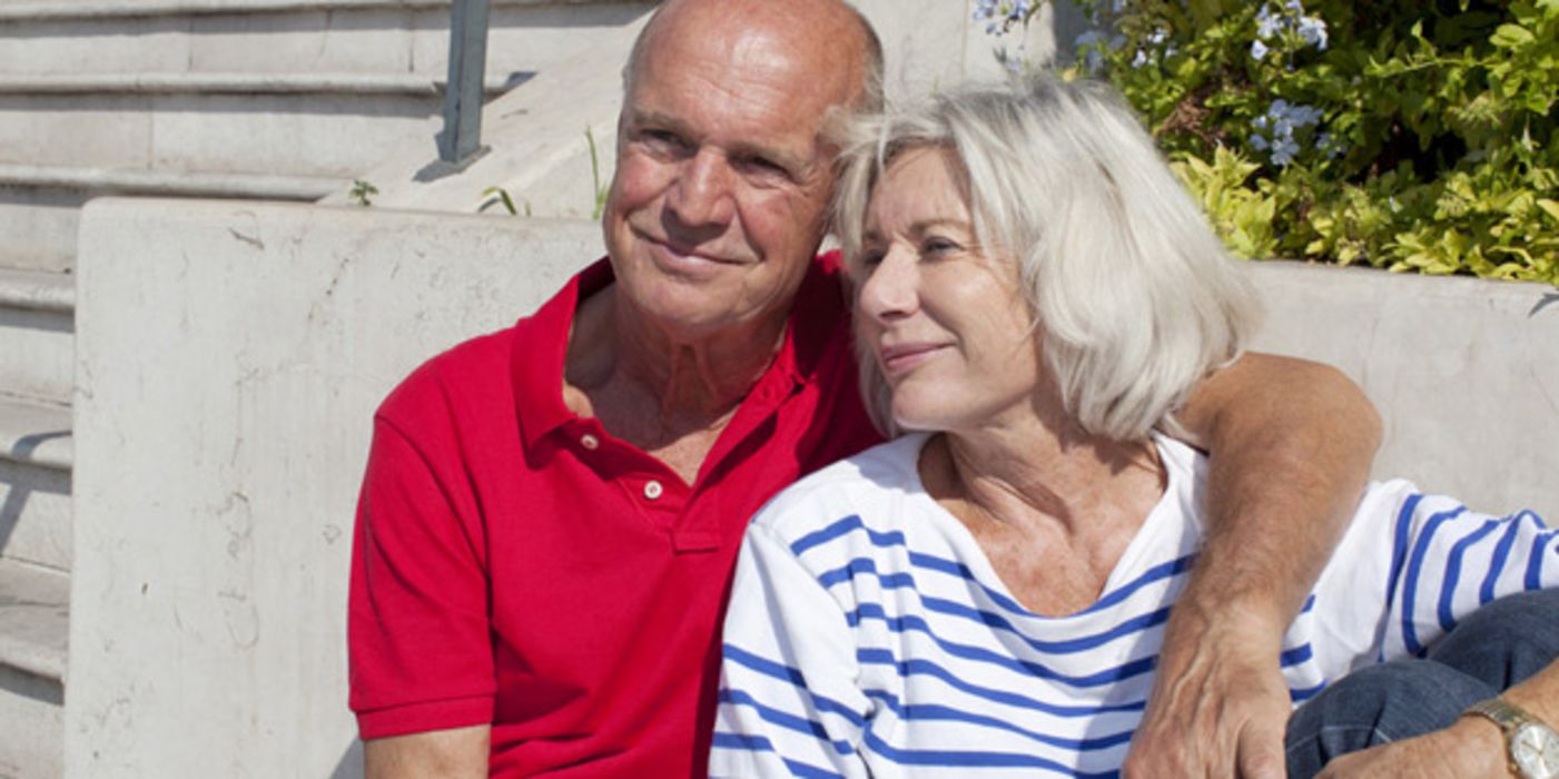 Älteres Paar sitzt in der Sonne; er im roten Polo-Shirt hat den Arm um ihre Schulter gelegt; sie trägt ein blau-weiß gestreiftes Top
