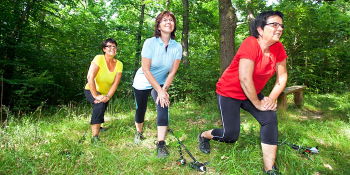 3 Frauen in Sportkleidung in den 50ern im Laubwald beim Dehnen, ein Paar Walkingstöcke am Boden